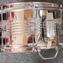 Stříbrný, chromovaný snare drum s kovovým korpusem. Na povrchu jsou vidět odrazy okolí a drobné nečistoty. Nástroj je vybaven ladicími šrouby a mecha…