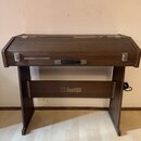 Hnědý klavír nebo digitální piano v dřevěném korpusu s uzavíracím víkem. Na přední spodní části je zlatý nápis „FAKFISA“ s doprovodným logem. Nástroj…