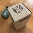 Na fotografii je bílá kartonová krabice a vedle ní tyrkysový efekt pedál. Na krabici je černý nápis TONE PRESS a PARALLEL COMPRESSOR, v dolní části j…