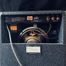 Zadní strana kytarového komba s černým texturovaným povrchem. V horní části jsou tři zlaté konektory s nápisy HEADPHONE, PREAMP OUT a REMOTE SWITCH,…