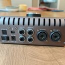 Zadní strana šedého audio rozhraní s kovovým povrchem a svislými ventilačními otvory v horní části. Na panelu jsou nápisy OPTICAL IN, POWER (s přepín…