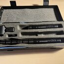 Černý kufřík s pěnovou výplní obsahuje dva mikrofony značky SENNHEISER a jeden černý plastový držák. Delší mikrofon má v přední části žebrovaný povrc…
