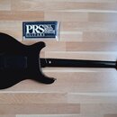 Zadní strana černé elektrické kytary s černým krytem elektroniky. Vedle hředu leží balení strun s nápisem PRS PAUL REED SMITH GUITARS. Nástroj je v č…