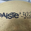 Detailní pohled na mosazný činel s viditelným rýhováním povrchu. Na čineli je černý nápis PAiSte 302 a pod ním označení 20" Ride. V levém dolním rohu…