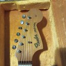 Hlava elektrické kytary z přírodního dřeva v otevřeném kufru s žlutým polstrováním. Na hlavě je černý nápis Fender JAGUAR a pod ním drobným písmem ne…