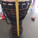 Fotografie zobrazuje djembe s černým korpusem zdobeným hnědými ornamenty. Nástroj má světlou membránu nataženou pomocí černého provázku. Přes nástroj…