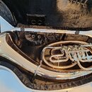 Stříbrný měděný nástroj (tuba) v černém polstrovaném kufru. Na povrchu jsou viditelné odrazy a drobné nečistoty. Nástroj má tři ventily a zakřivené t…