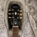 Černá kytara v otevřeném hnědém plišovém pouzdře. Na hlavě nástroje je bílý nápis Epiphone a pod ním bílé logo v podobě stylizedovaného písmene „E“.…