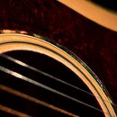 Detailní záběr na soundhole akustické kytary. Okraj otvoru je zdoben vícevrstvým hnědým bindingem a tmavě červeným vzorníkem s mramorovým efektem. Na…