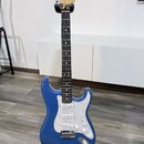 Modrá elektrická kytara s třemi snímači a bílým vzorovaným pickguardem, umístěná na černém stojanu. Na hlavě hředu je viditelné logo Fender. Kytara m…