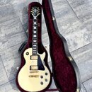 Kytara typu electric guitar v krémové barvě s černým pickguardem, umístěná v otevřeném tvrdém kufru s vínovým polstrováním. Na hlavě hmatníku je vidi…