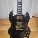 Černá elektrická kytara s dvojstupňovitým tělem a zlatým hardwarem. Na hmatníku jsou vidět bílé vložky a na těle jsou dva zlaté humbuckery, zlatý mos…