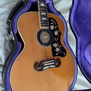 Akustická kytara s přirozeným dřevěným povrchem v černém tvrdém kufru s fialovým vnitřkem. Nástroj má černý pickguard s květinovým ornamentem a černý…