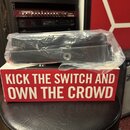Černý efekt pedál zabalený v průhledné plastové fólii. Pedál leží na červeném boxu s bílým nápisem KICK THE SWITCH AND OWN THE CROWD. V pozadí je vid…