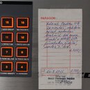 Fotografie zachycuje detail ovládacího panelu s oranžovými podsvícenými tlačítky s čísly 15, 16, 11, 12, 7, 8, 3 a 4. Vedle panelu je položen papírov…