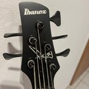 Černá hlava basové kytary s bílým logem Ibanez. Na povrchu je viditelný bílý podpis, který je částečně nečitelný. Jsou zde vidět čtyři ladičky a stru…