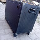 Černý transportní case s koly, vyrobený z dřevěného materiálu s tmavým povrchem. Case má kovové hrany, vrchy a madla. Povrch vykazuje známky opotřebe…