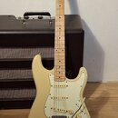 Krémová elektrická kytara typu Stratocaster s bílým pickguardem a javorovým hmatníkem. Na hlavě je viditelné logo Squier. Kytara je umístěna před čer…