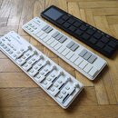 Tři bílé a černé MIDI kontroléry značky KORG položené na dřevěné podlaze. První je bílý kontrolér s posuvníky a tlačítky s nápisem nanoKONTROL2. Pros…