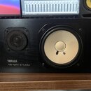 Černý studiový monitor s bílým membránovým reproduktorem a menším výškem krytým černou mřížkou. V levém dolním rohu je bílý nápis YAMAHA a pod ním NS…