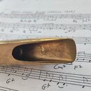 Detailní pohled na hutnou, mosaznou dutinu mouthpiece s viditelným opotřebením a drobnými škrábanci vnitřního povrchu. Předmět leží na listu s hudebn…