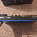 Modrá kytarový procesor s bílým nápisem BOSS GT-1. Na zadní straně jsou vstupy a výstupy s popisky INPUT, AUX IN, PHONES, OUTPUT R, L/MONO a CTL2.3/E…