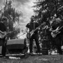 Černobílá fotografie zachycuje tři hudebníky s nástroji na venkovním pódiu. První osoba zleva hraje na světlou elektrickou kytaru s dvěma humbuckery,…