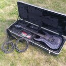 V otevřeném černém kufru s polstrováním leží elektrická kytara s tmavě fialovým nebo šedým vzorným povrchem těla. Vedle kytary jsou v kufru umístěny…