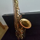 Zlatý saxofon s kovovým povrchem, který leží v otevřeném černém kufru. Nástroj má v horní části tmavý nástavec a v dolní části je přiložen samostatný…