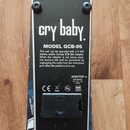 Zadní strana efektového pedálu v černé barvě. Na povrchu je bílý nápis cry baby, MODEL GCB-95 a text s instrukcemi k napájení. V dolní části je logo…