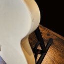 Detail zadní strany bílé elektrické kytary s lesklým povrchem. Nástroj je umístěn v černém stojanu na dřevěném stole. Na hraně korpusu je viditelný d…