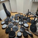 Sada komponentů elektronické bicí soupravy rozložená na dřevěné podlaze. Je zde vidět několik padů různých velikostí, černé gumové činely a kovový rá…