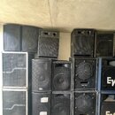 Sestava různých černých reproduktorových beden v několika řadách. Na některých jsou vidět loga EV a nápisy BASS 150 S. Povrchy některých beden vykazu…