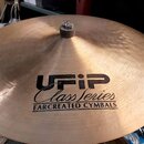 Zlatavý kovový činel s viditelným broušením povrchu. Uprostřed je černý nápis „UFIP Class Series EARCREATED CYMBALS“. V centru činelu je umístěn tlum…