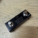 Černý kovový externí spínáč s dvěma stříbrnými tlačítky. Na horní straně je bílý nápis AMPERO SWITCH a pod ním menší text Momentary Footswitch. Na pr…