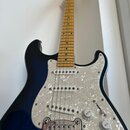 Elektrická kytara s tmavě modrým průsvitným lakem, přes který je vidět struktura dřeva. Nástroj má bílý perleťový pickguard, tři snímače s bílými pla…