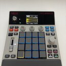 Šedý sampler MPC SAMPLE značky AKAI professional. Zařízení má v horní části displej s vizualizací zvukové vlny, ovládací knoflíky a v pravé části 16…