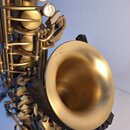 Detailní pohled na saxofon v matném zlatém provedení. Jsou vidět klapy s perleťovými vložkami a mechanismus nástroje. Na povrchu kónického zvonu jsou…