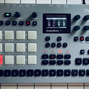 Šedý analogový drum computer a sampler značky Elektron, model Analog Rytm MKII. Zařízení má v horní části řadu ovládacích knoflíků, uprostřed displej…