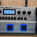Stříbrný basový efektový procesor s černým pedálem. Na horní části je nápis BOSS GT-1B BASS EFFECTS PROCESSOR, displej a několik ovládacích knoflíků…