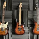 Tři elektrické kytary s hnědým plamenovitým povrchem těla zavěšené na šedém akustickém panelu. Nejlevější kytara vlevo má bílý pickguard a na hlavě l…