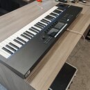 Černý MIDI kontrolér s klávesnicí položený na dřevěném stole. Na pravé straně jsou umístěny kontrolní prvky, padsy a displej, v pravém horním rohu je…