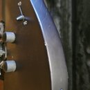 Detailní pohled na tmavě hnědou kytaru s matným povrchem. Na těle jsou vidět dva černé oválné knofíky a jeden přepínač s kovovou páčkou. V dolní část…