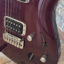 Detail elektrické kytary v tmavě červené barvě s lesklým povrchem. Nástroj má dva humbuckery s chromovanými rámečky, chromovaný most a dvě ovládací k…