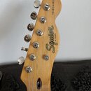Fotografie zachycuje hlavu elektrické kytary z přírodního světlého dřeva. Na povrchu je černý nápis Squier BY FENDER a svisle orientovaný nápis TELEC…