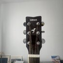 Fotografie zachycuje hlavu akustické kytary v černé barvě. V horní části je bílý nápis YAMAHA a pod ním bílé logo ve tvaru stylizovaného ptáka. Nástr…
