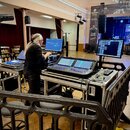 Fotografie zachycuje zvukovou režii v sále. Na dvou černých flightcasech jsou umístěny digitální mixážní pulty a dva počítačové monitory. Za pultem s…