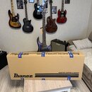 Na fotografii je v popředí velká hnědá papírová krabice s nápisem Ibanez a ELECTRIC GUITARS & BASSES, která leží na bílo-hnědém stolku. Za krabicí st…