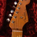 Detail hlavy dřevěné elektrické kytary v červeném plišovém pouzdře. Na dřevě je vidět nápis "Fender" a nad ním svisle text "STRATOCASTER". V horní čá…