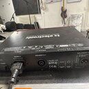 Černý rackový zesilovač s červeným horním profilem. Na horní straně je bílý nápis tc electronic a text DESIGNED AND ENGINEERED IN DENMARK. Na zadním…